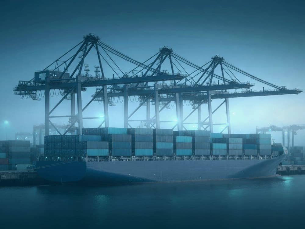 Container ship berthed at a marine terminal at dusk, with gantry cranes working under floodlights.